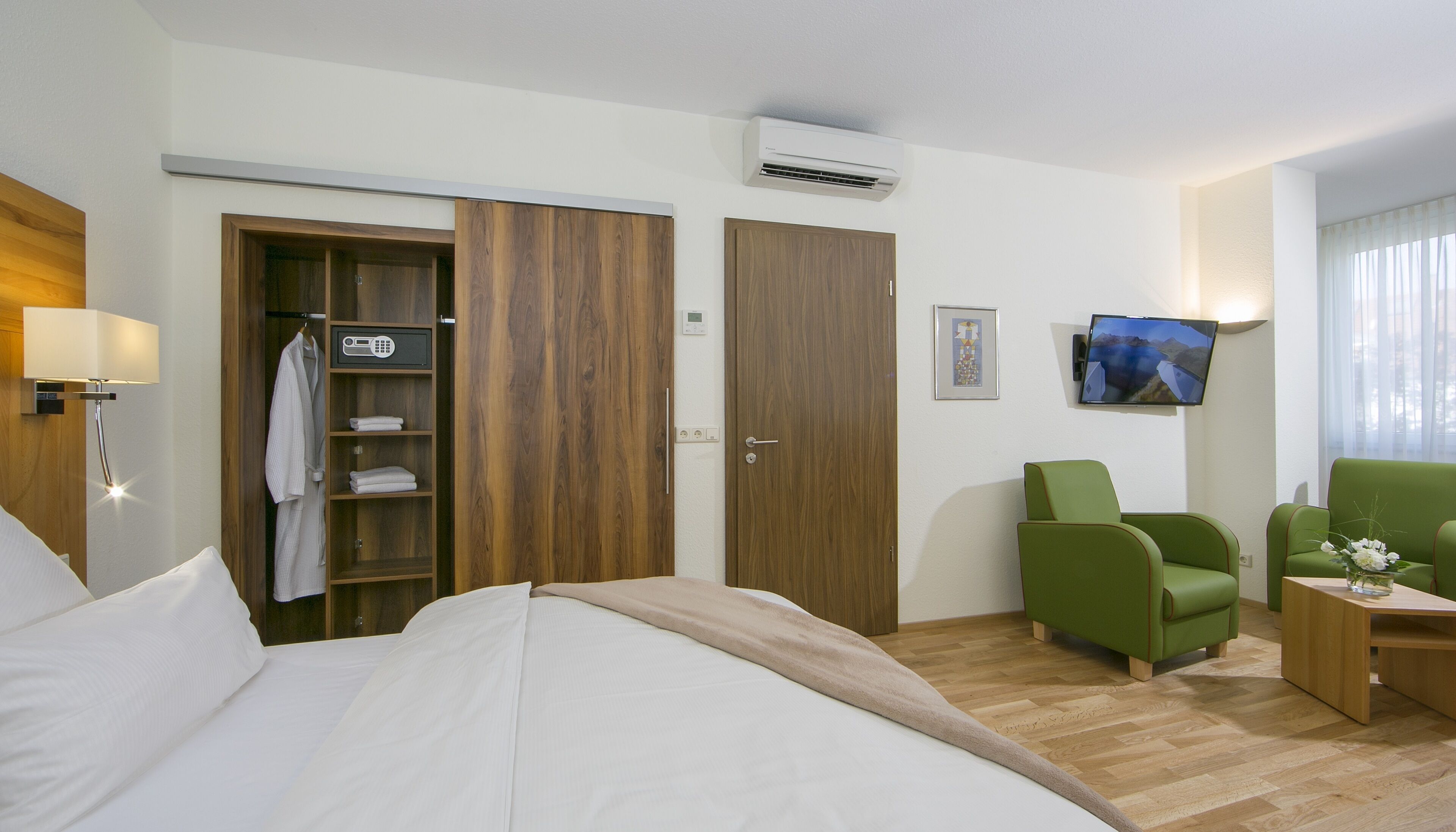 Habitación de hotel con una cama, un armario, un sillón verde, una mesita y un televisor en la pared.