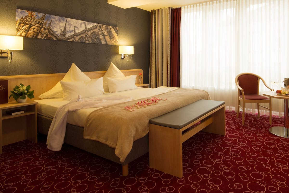 A hotel room with a bed, bedside tables, a desk, chairs, and a large window with curtains.