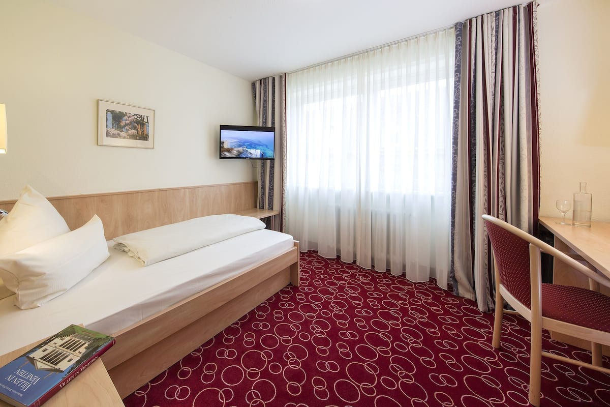 A hotel room with a bed, a TV, a desk, a chair, a lamp, a window with curtains, and a carpet.