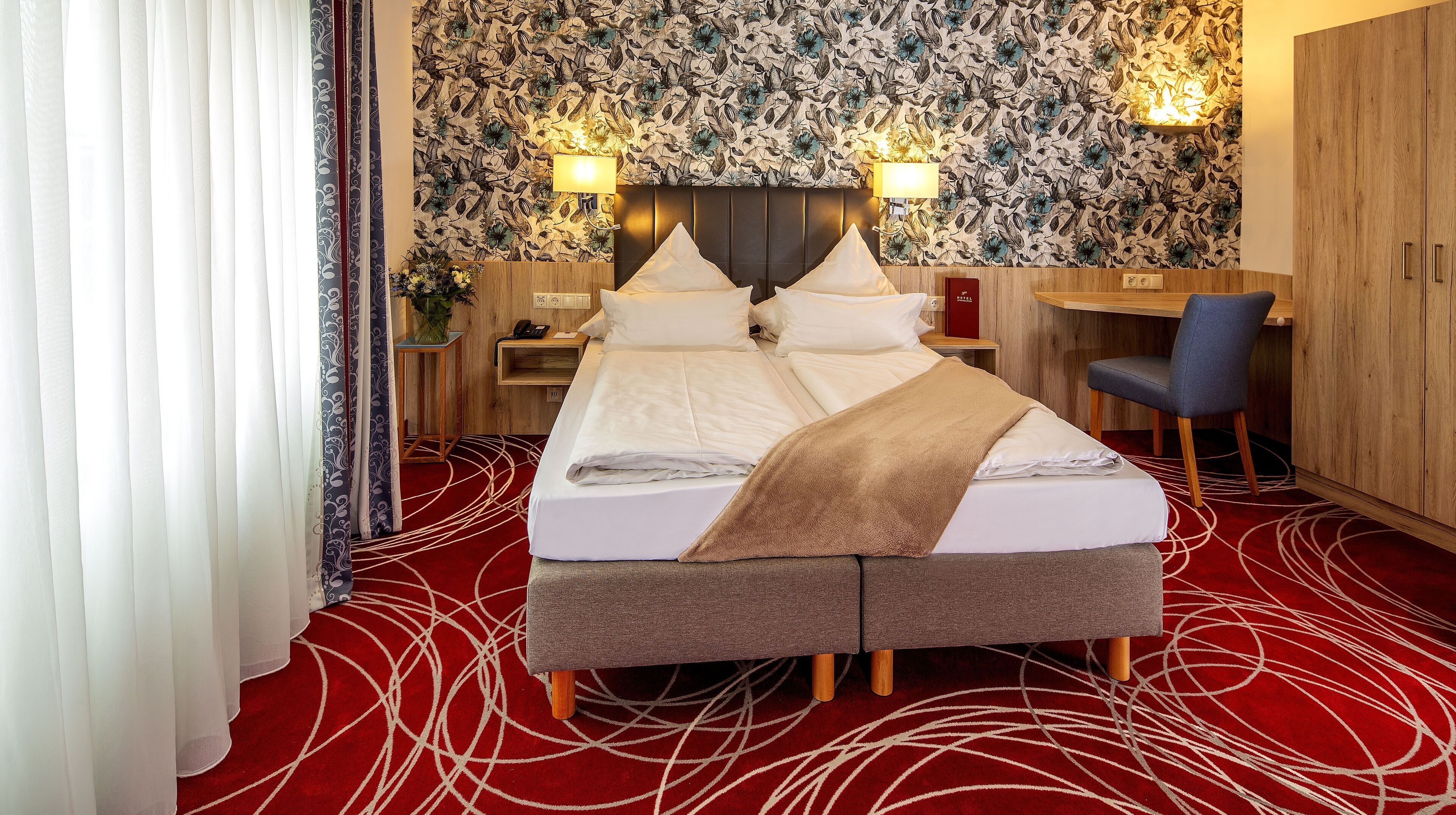 A hotel room with a large bed, a desk, and a chair. There are two bedside tables with lamps. The room has a floral wallpaper and a red carpet.