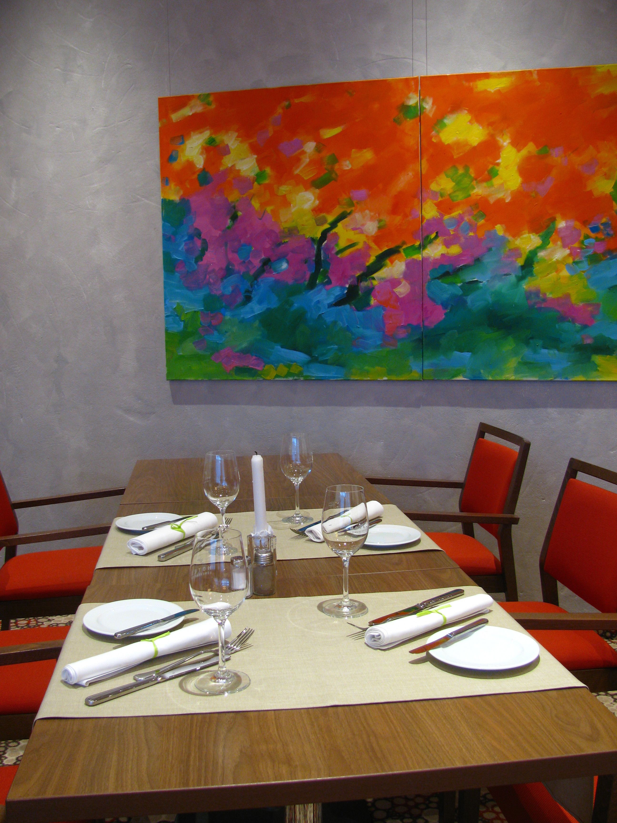Uma mesa de jantar posta para quatro pessoas, cadeiras vermelhas, um quadro abstrato colorido na parede e uma mesa de madeira.