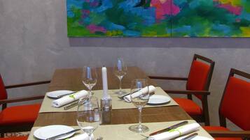 Uma mesa de jantar posta para quatro pessoas, cadeiras vermelhas, um quadro abstrato colorido na parede e uma mesa de madeira.
