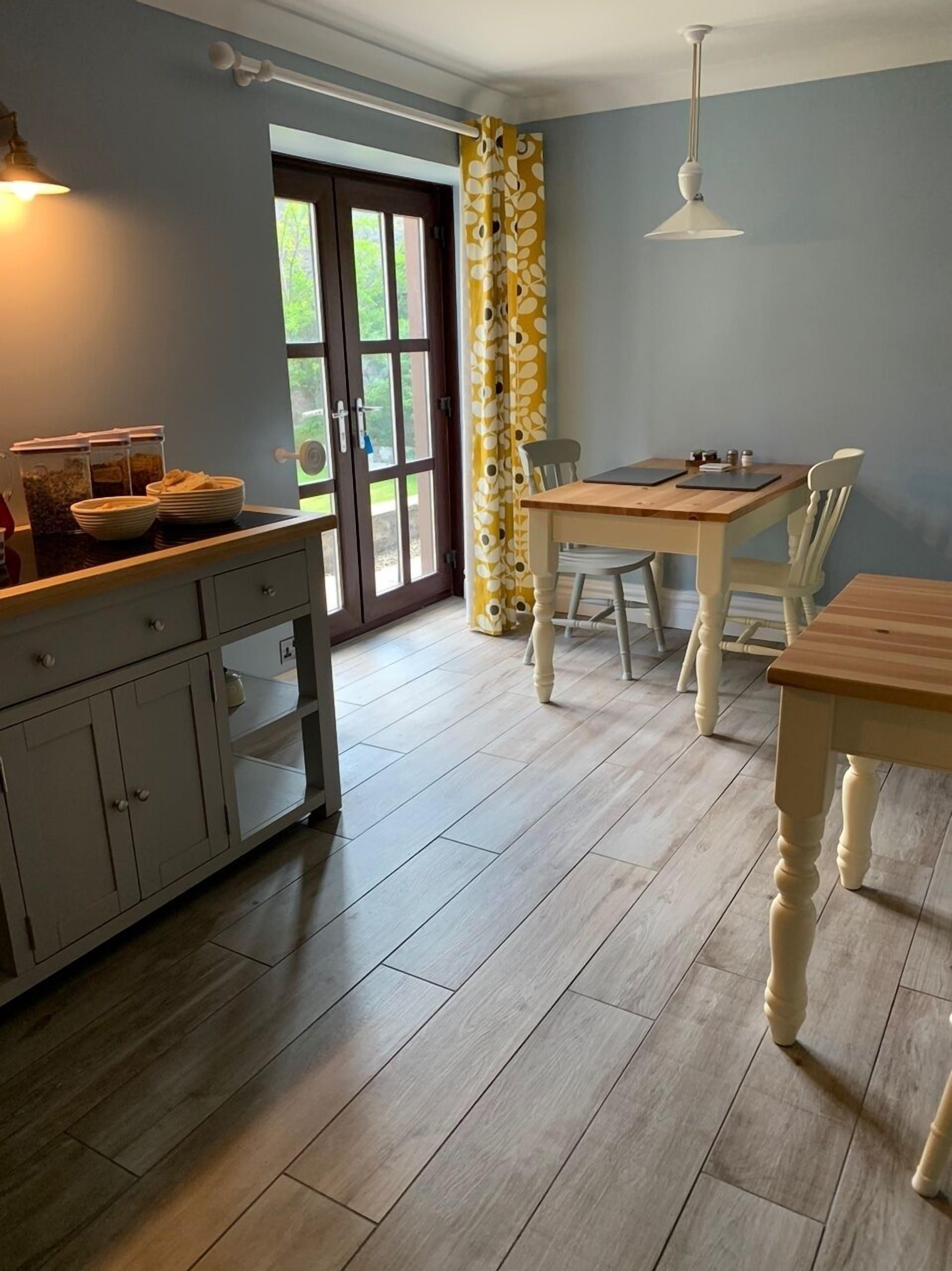 A kitchen with a wooden table and chairs, a cabinet with drawers, a window with curtains, and a door leading outside.