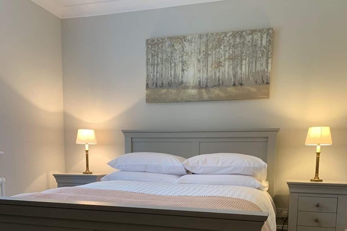 A bedroom with a bed, two bedside tables with lamps, a nightstand, a wall-mounted painting, and a wooden floor.