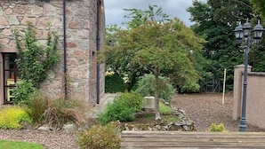 Un bâtiment en pierre doté d’un toit en pente, entouré d’un jardin où poussent différentes espèces de plantes et agrémenté d’un banc en bois.