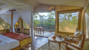 A bedroom with a bed, a bedside table, a chair, and a window with a view of trees. - Mara Leisure Camp (Maasai Mara)