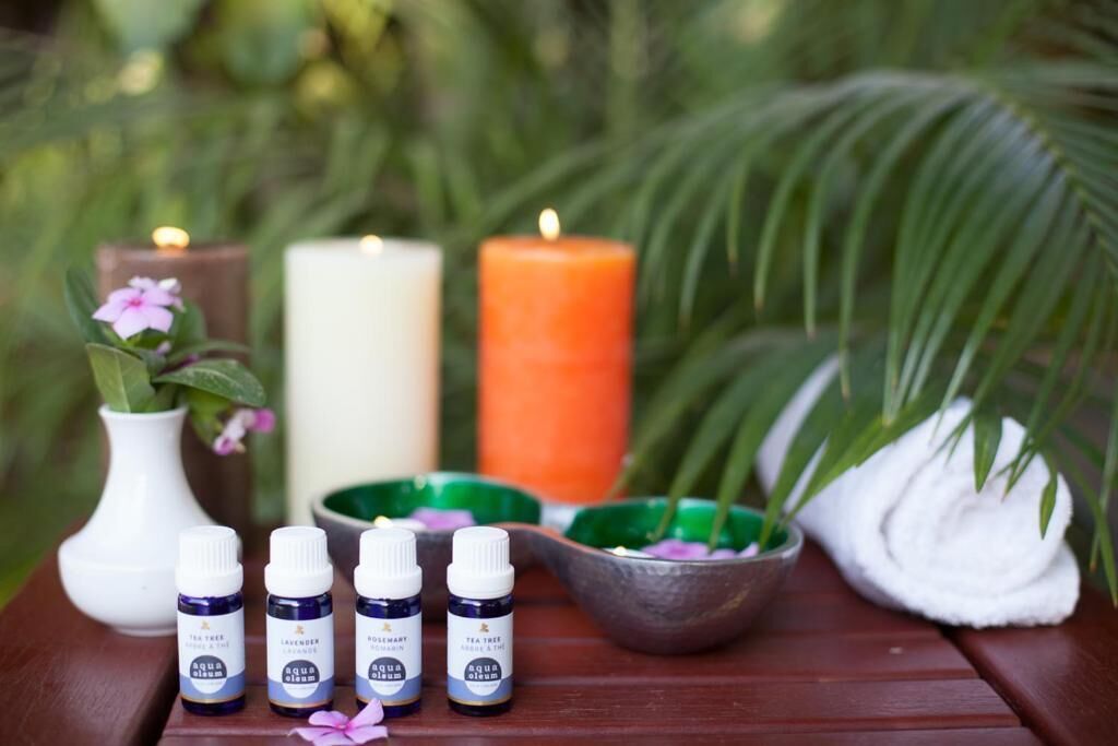 Uma mesa de madeira com quatro frascos de óleo de melaleuca, um vaso branco com uma flor rosa, duas velas, uma tigela com folhas verdes e uma toalha branca.