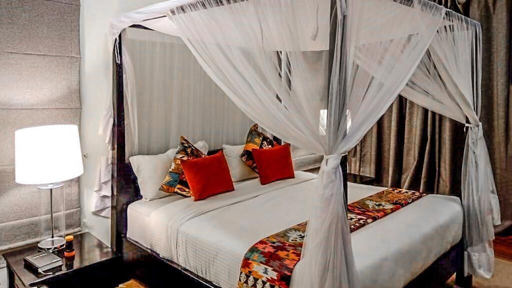 A four-poster canopy bed with white linens and colorful throw pillows, a bedside table with a lamp, and a view of the room's interior.