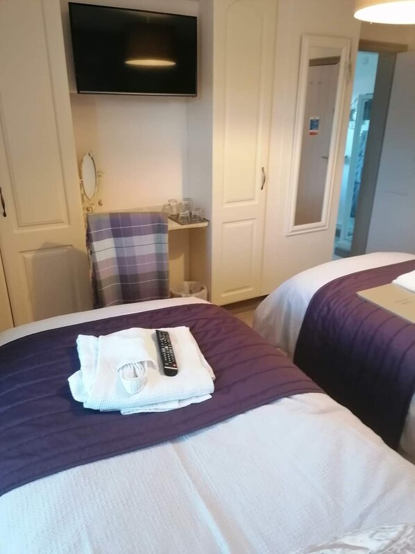 Habitación de hotel con dos camas, un televisor montado en la pared, una mesita con una silla y una puerta que da a otra habitación.