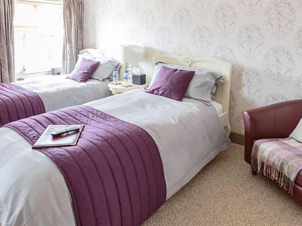 A hotel room with two beds, each with a purple and white bedspread, a remote control on the left bed, and a red armchair with a patterned throw.
