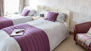 A hotel room with two beds, each with a purple and white bedspread, a remote control on the left bed, and a red armchair with a patterned throw.