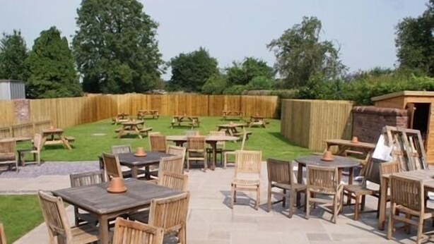 An outdoor patio area with stone flooring, wooden tables and chairs, and a wooden fence in the background. - The Sun Inn (Ellesmere)