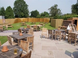 An outdoor patio area with stone flooring, wooden tables and chairs, and a wooden fence in the background. - The Sun Inn (Ellesmere)