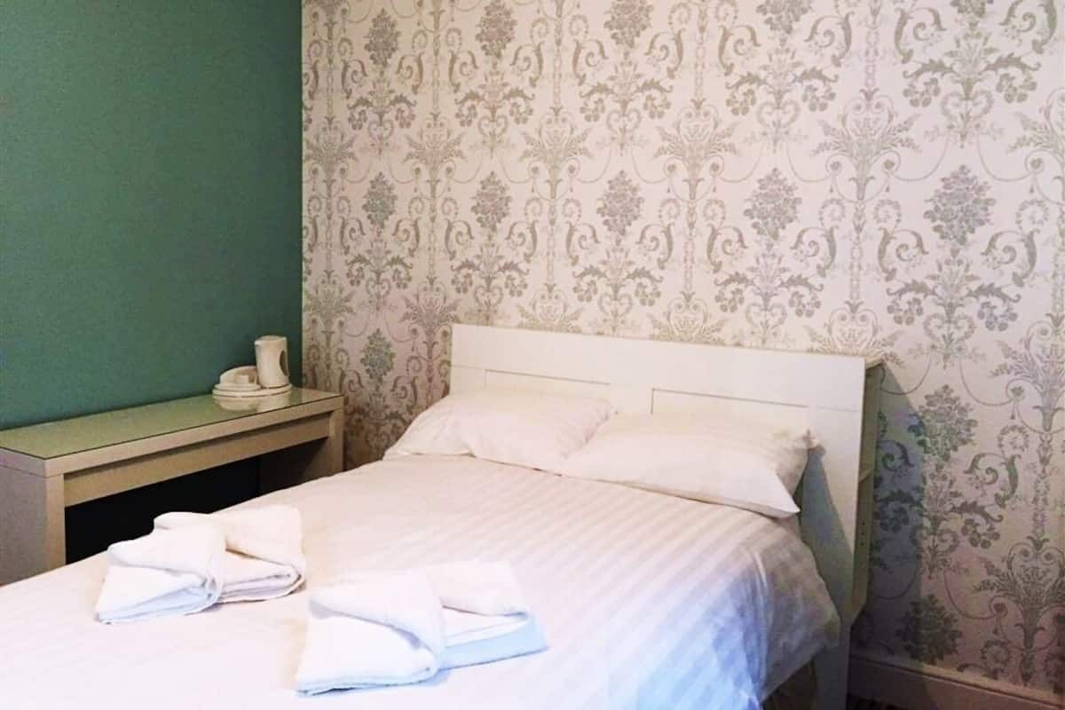 Cama bem arrumada com lençóis e almofadas brancos, mesa de cabeceira com uma xícara e um prato, e parede com papel de parede estampado com flores.