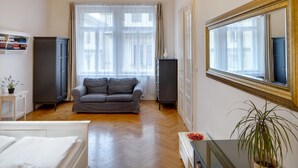 Un saló modern amb un sofà gris, un llit blanc, un paviment de fusta, una làmpada penjant i un mirall gran.