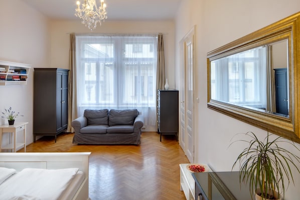 A modern living room with a grey sofa, a white bed, a wooden floor, a chandelier, and a large mirror.
