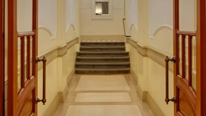 Un passadís ample amb escala, portes de fusta decorades i aplics de paret.