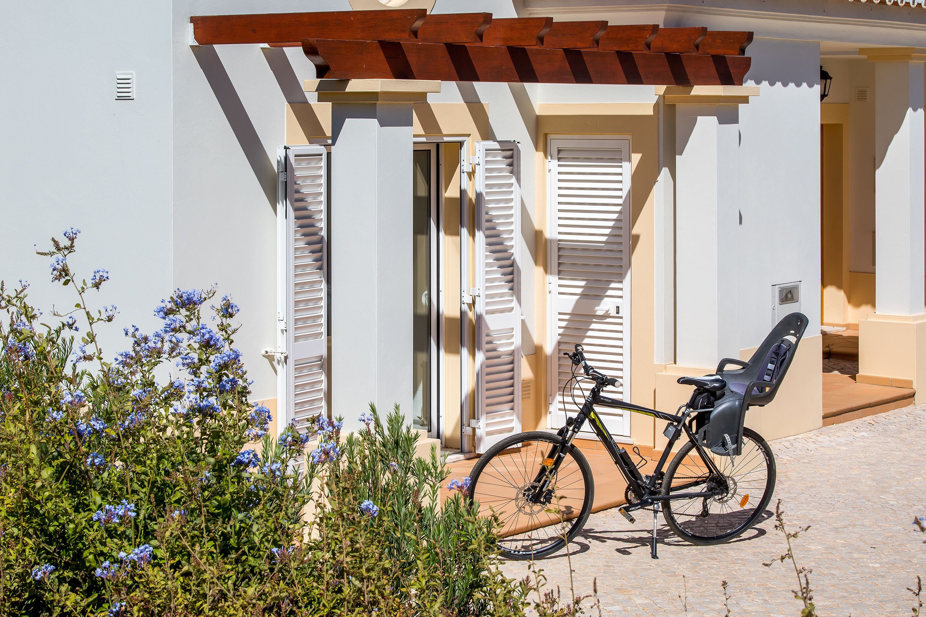 A bicycle parked outside a building with white shutters and a wooden pergola.