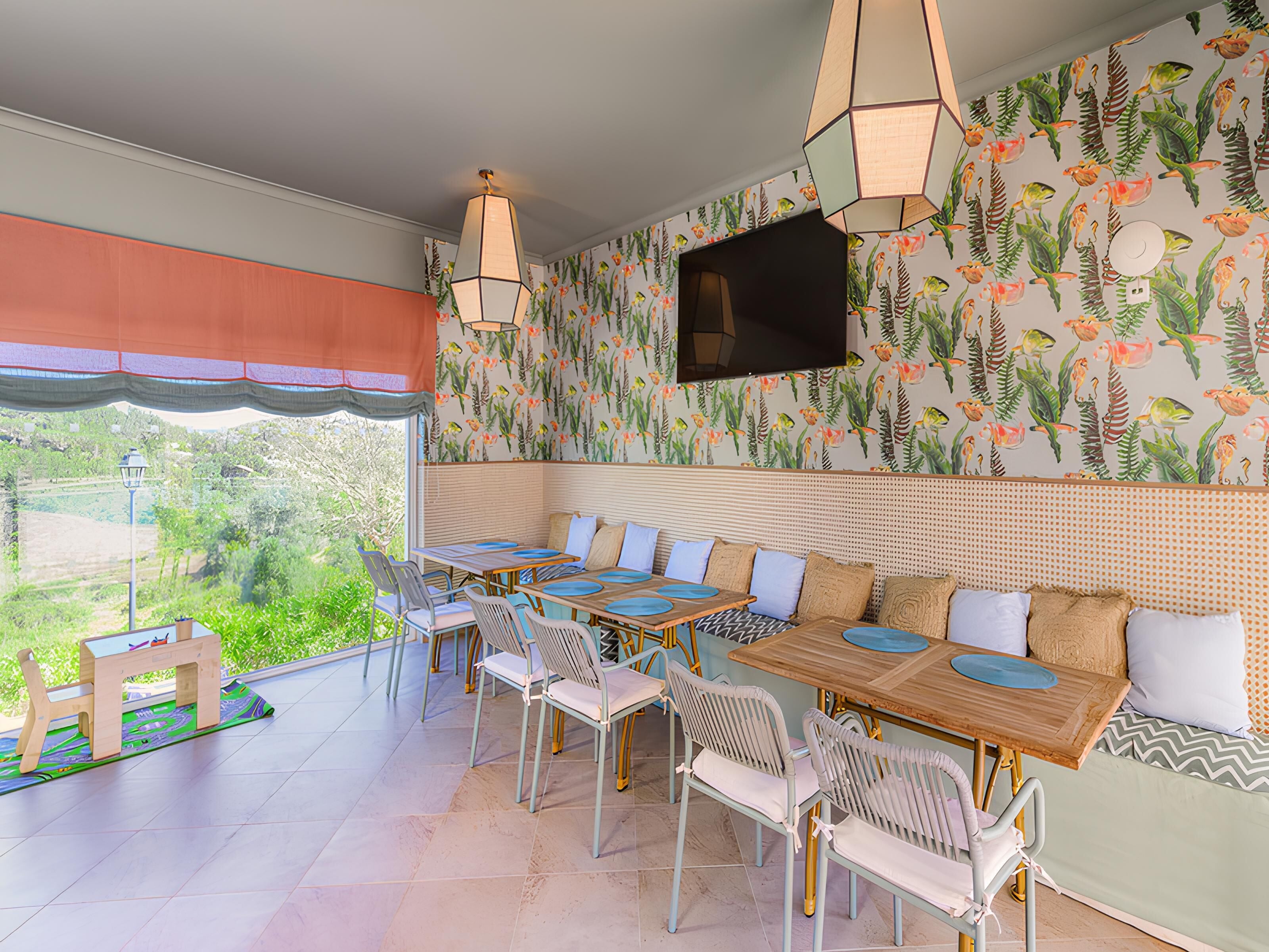 A dining area with a view of greenery outside, a TV mounted on the wall, and a patterned wallpaper.