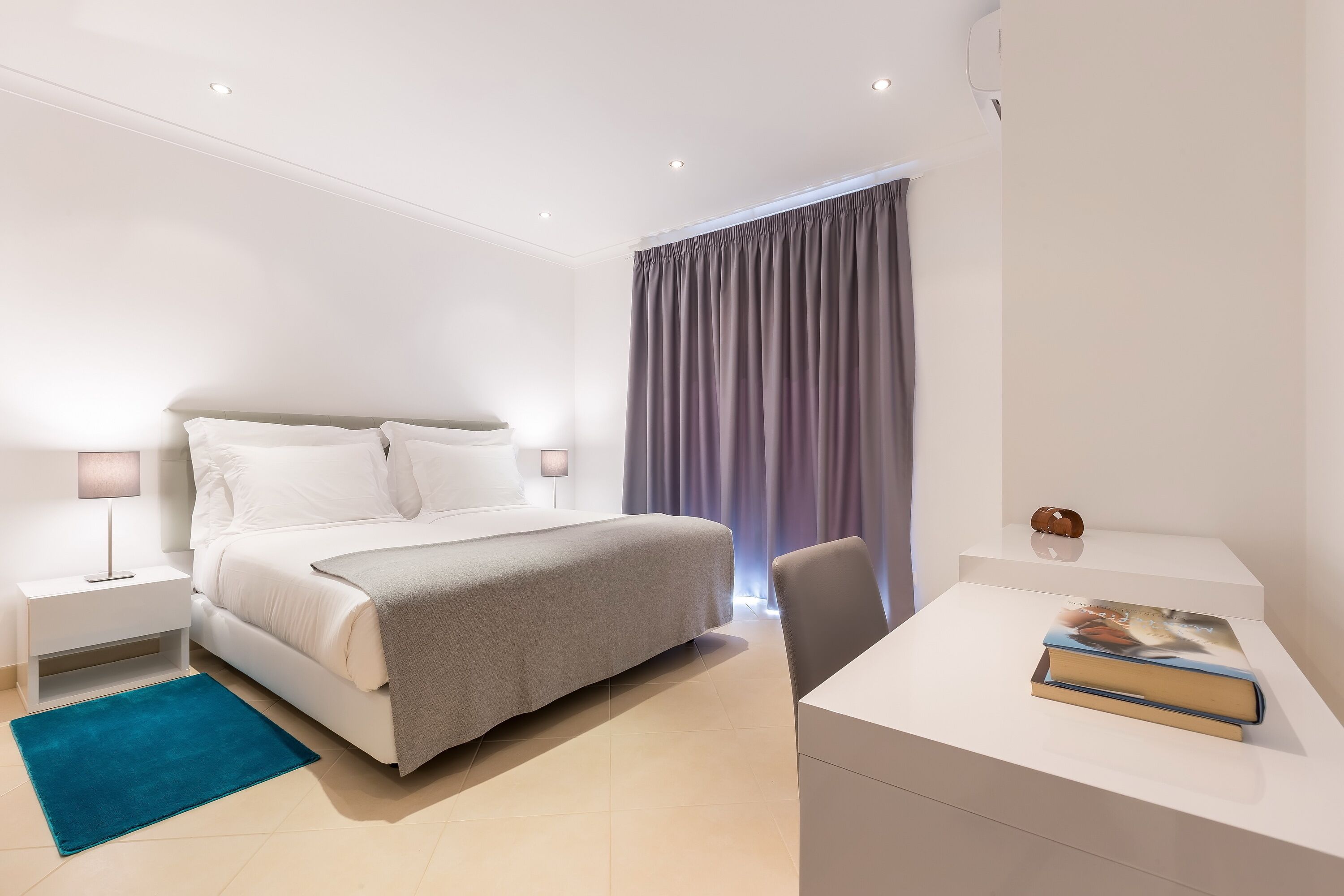 A modern hotel room with a neatly made bed, a desk with a book, and a blue rug.