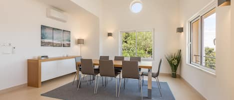 A modern dining area with a wooden table, six chairs, a grey rug, a white cabinet, a painting, a potted plant, and a window with a view of greenery.