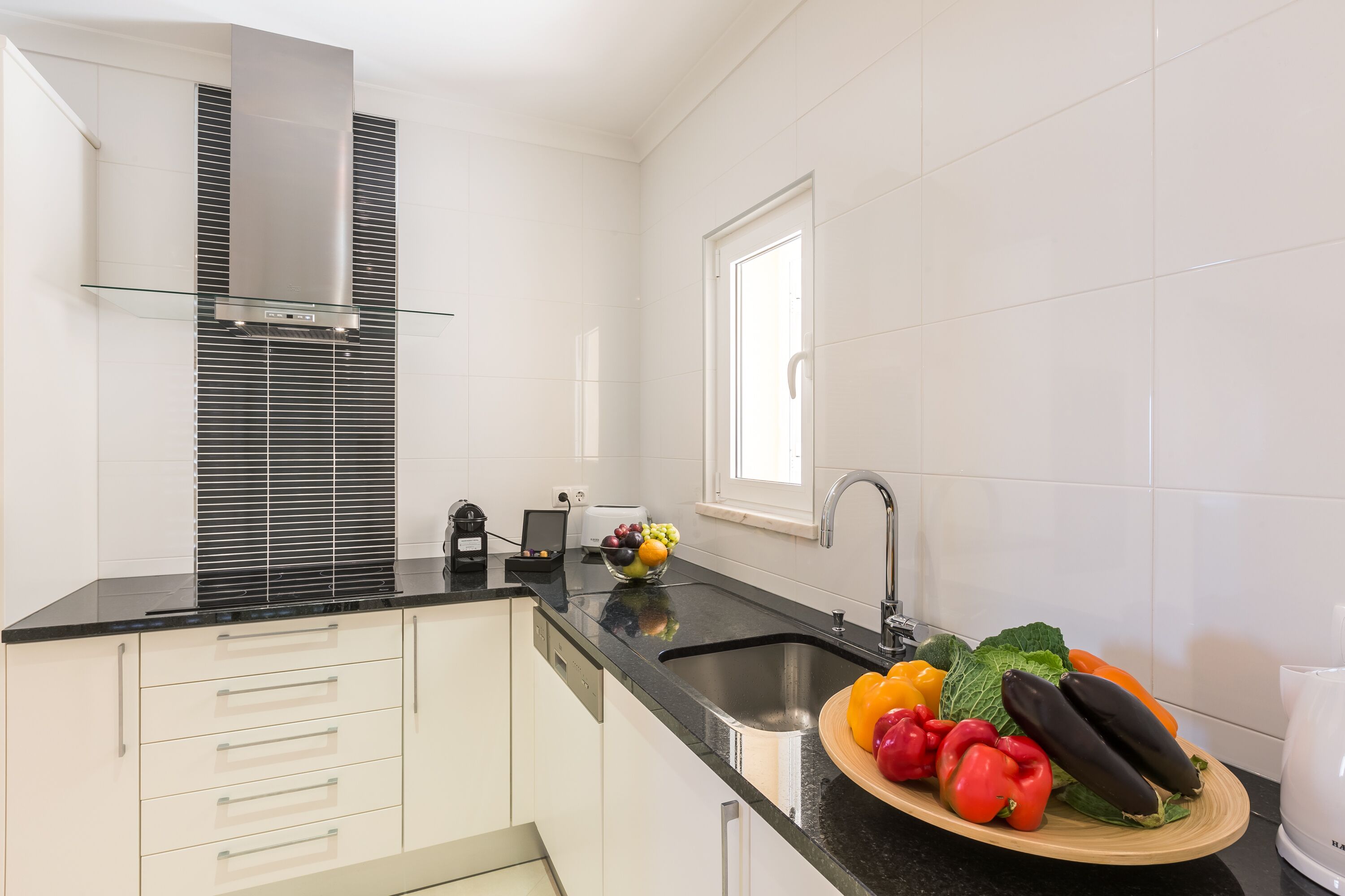 A modern kitchen with white cabinets, a black countertop, a stainless steel range hood, a sink, and a bowl of fresh vegetables.