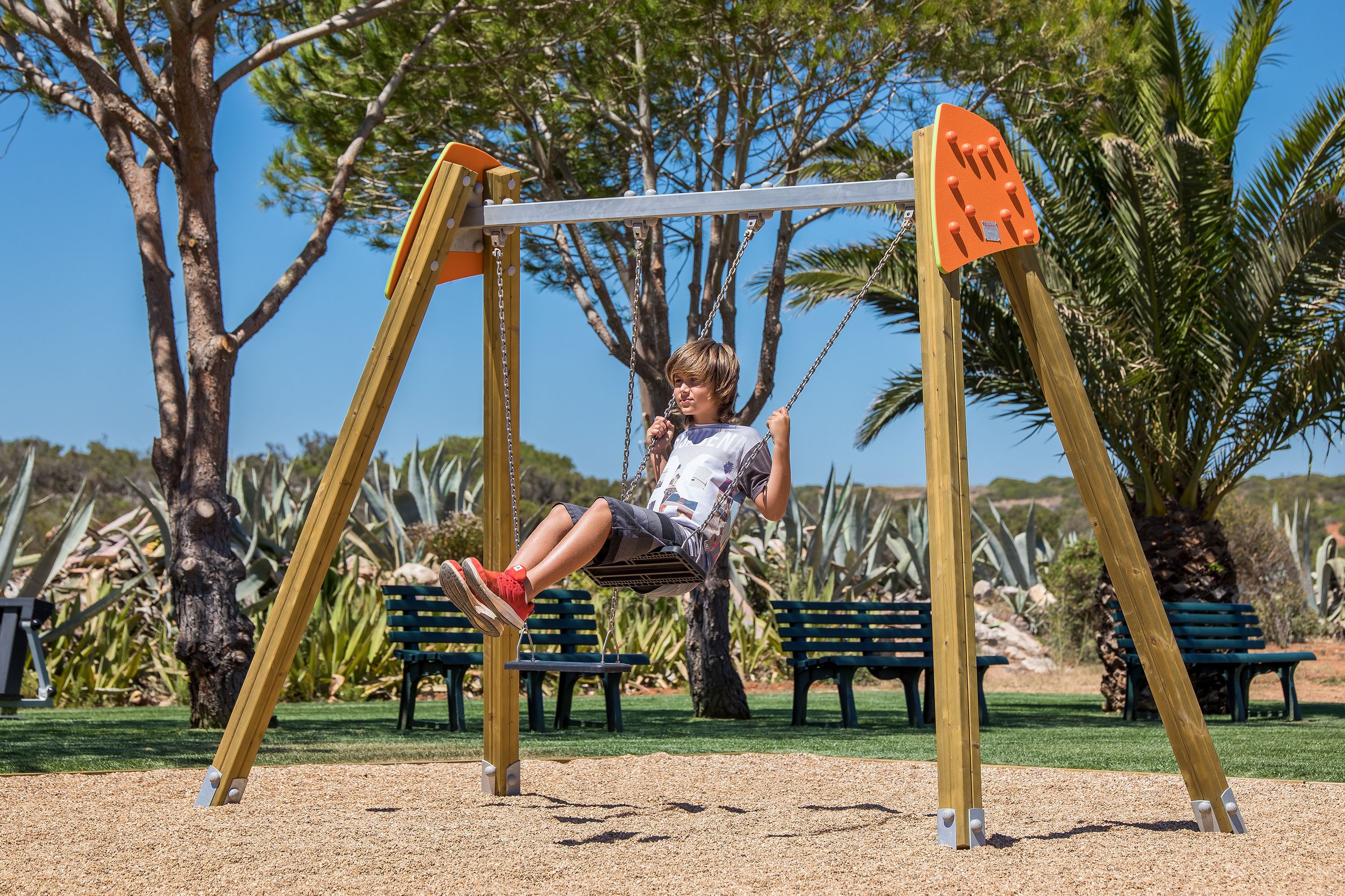 A swing set in a park with benches and palm trees in the background.