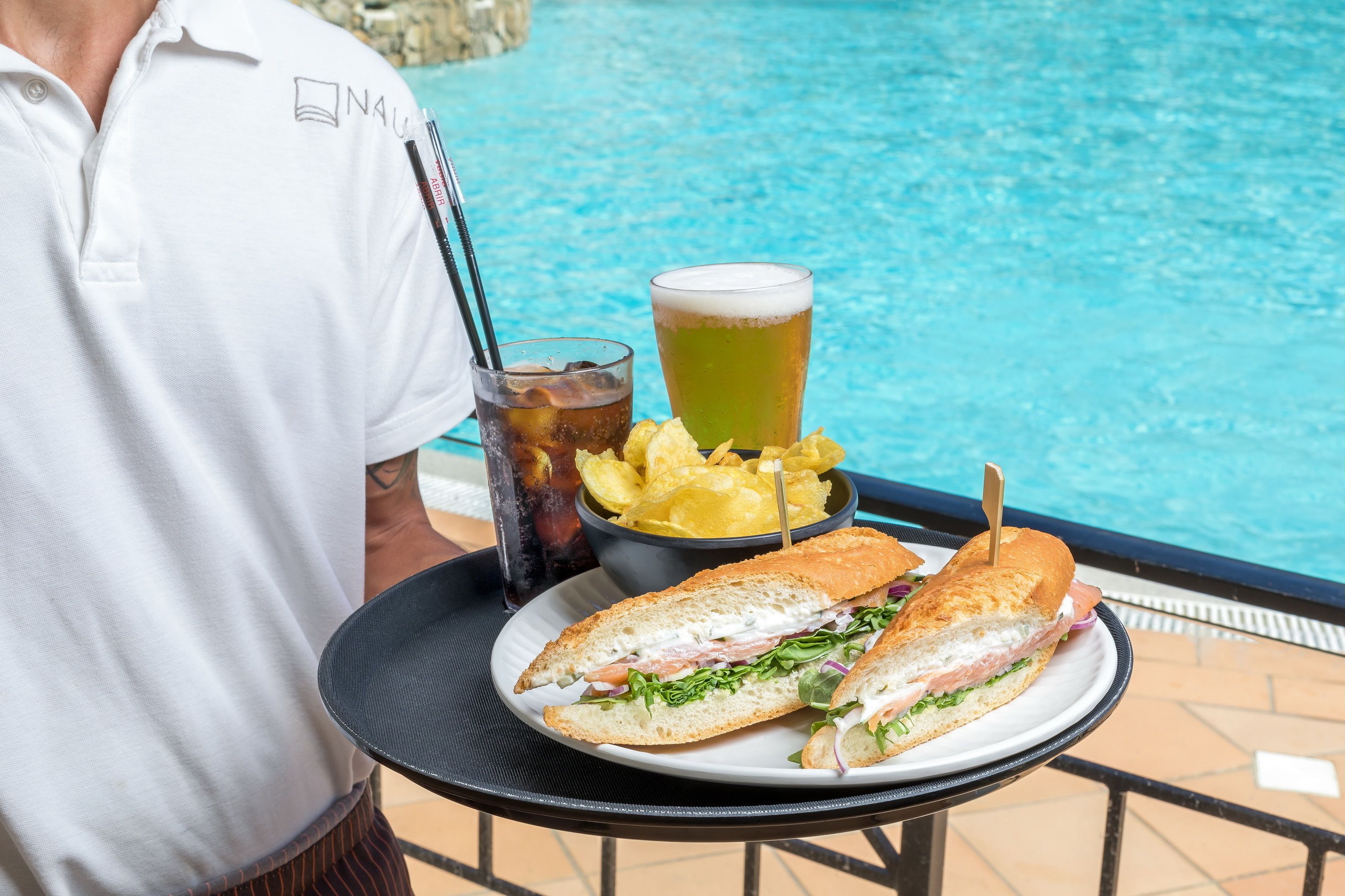 A tray with a sandwich, chips, a drink, and a glass of beer by a poolside.