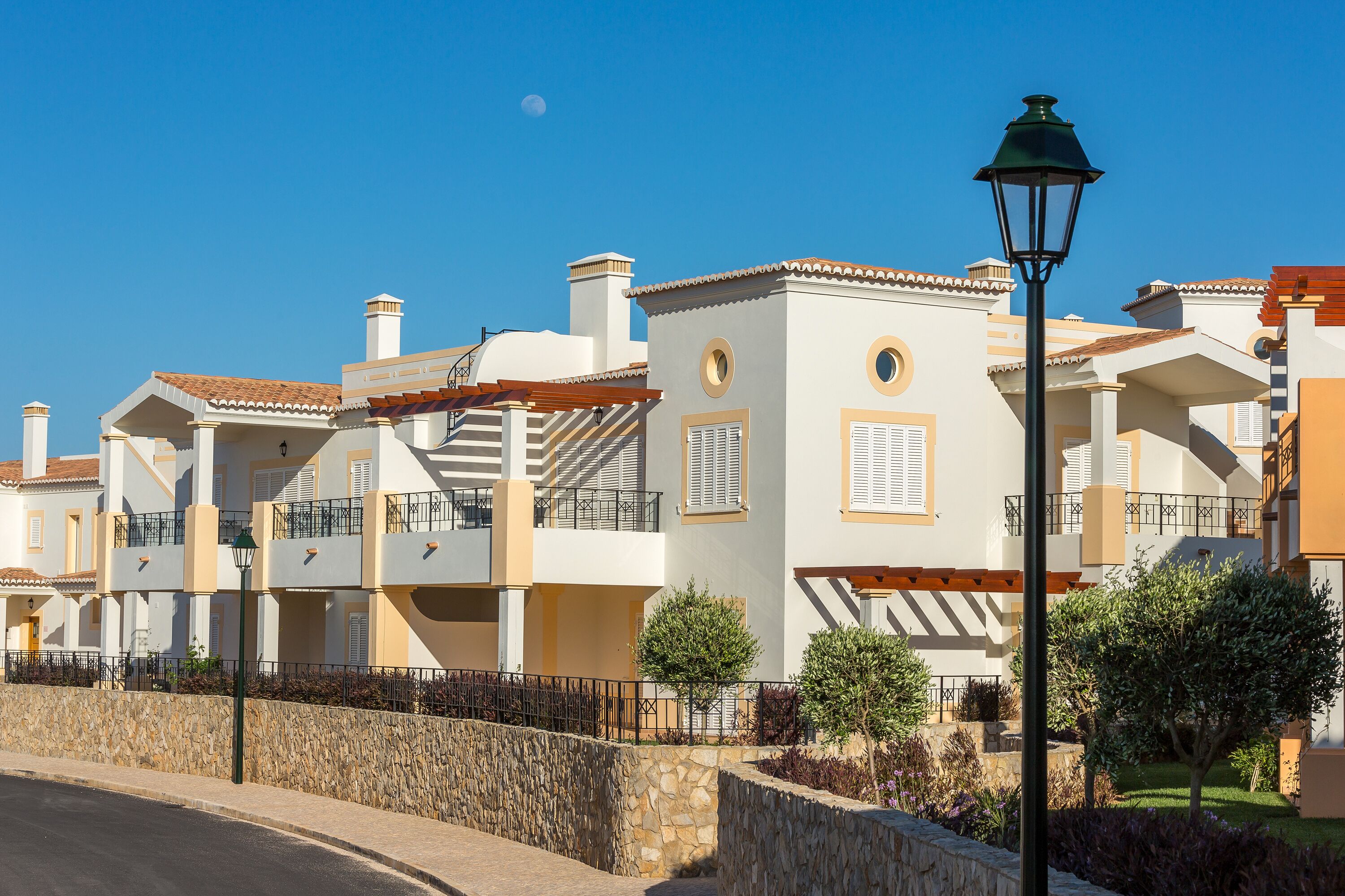 A row of modern, two-story buildings with balconies, a stone wall, and a street lamp.