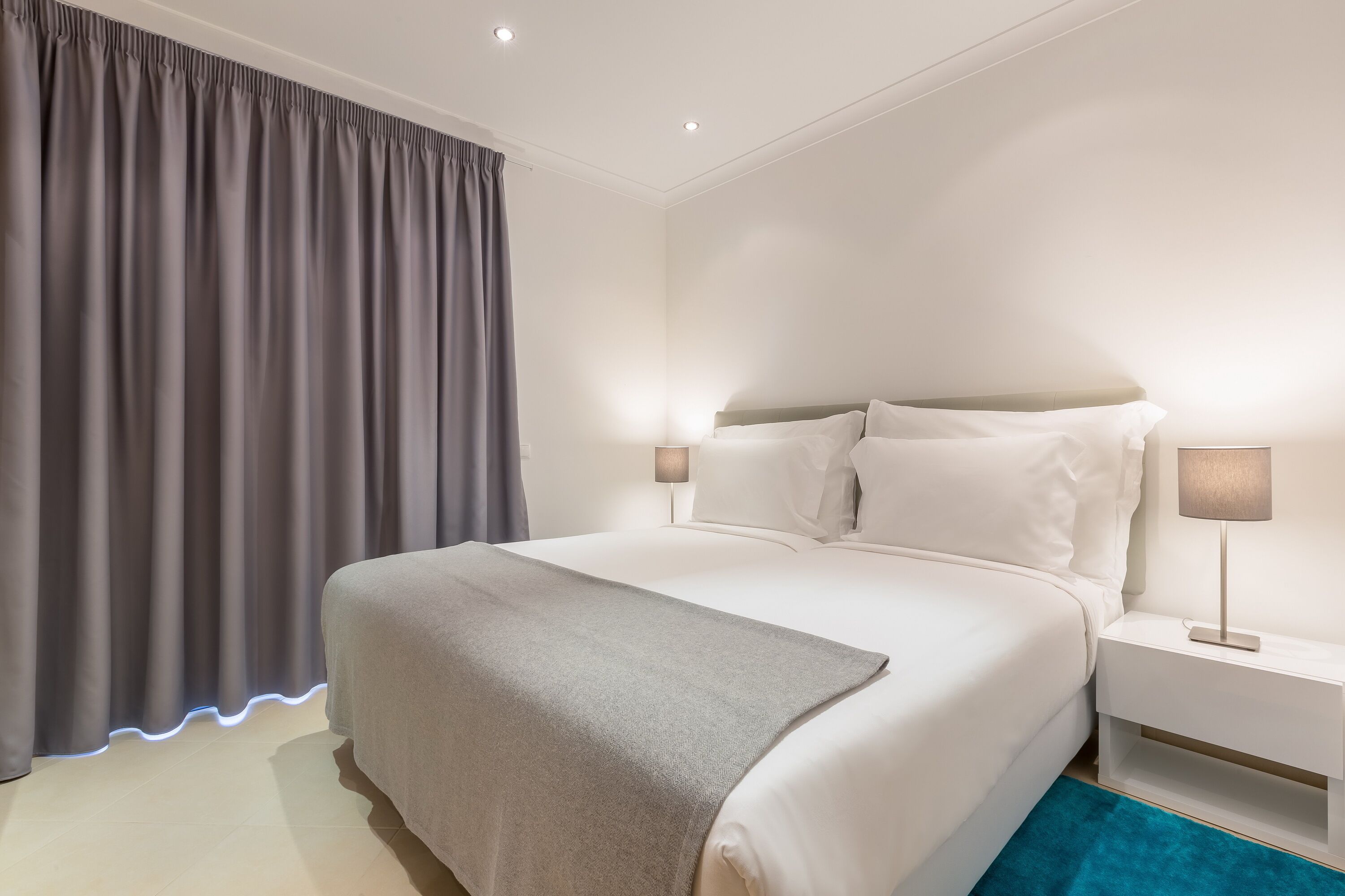 A neatly made bed with a grey blanket, a white bedside table with a lamp, and a grey curtain.