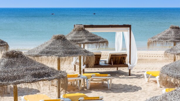 Een strand met rieten parasols, ligbedden en een zonnescherm.