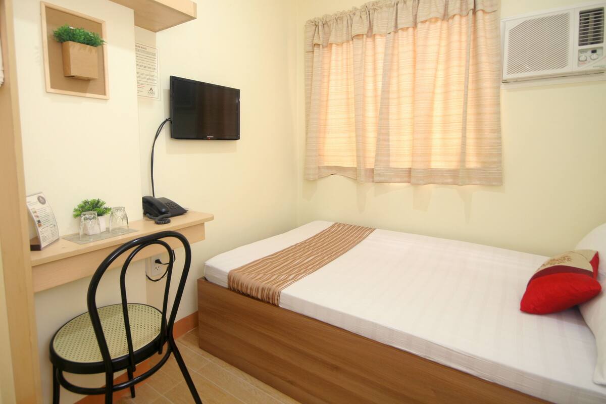 A bedroom with a bed, a desk with a computer, a chair, a TV, and a window with curtains.