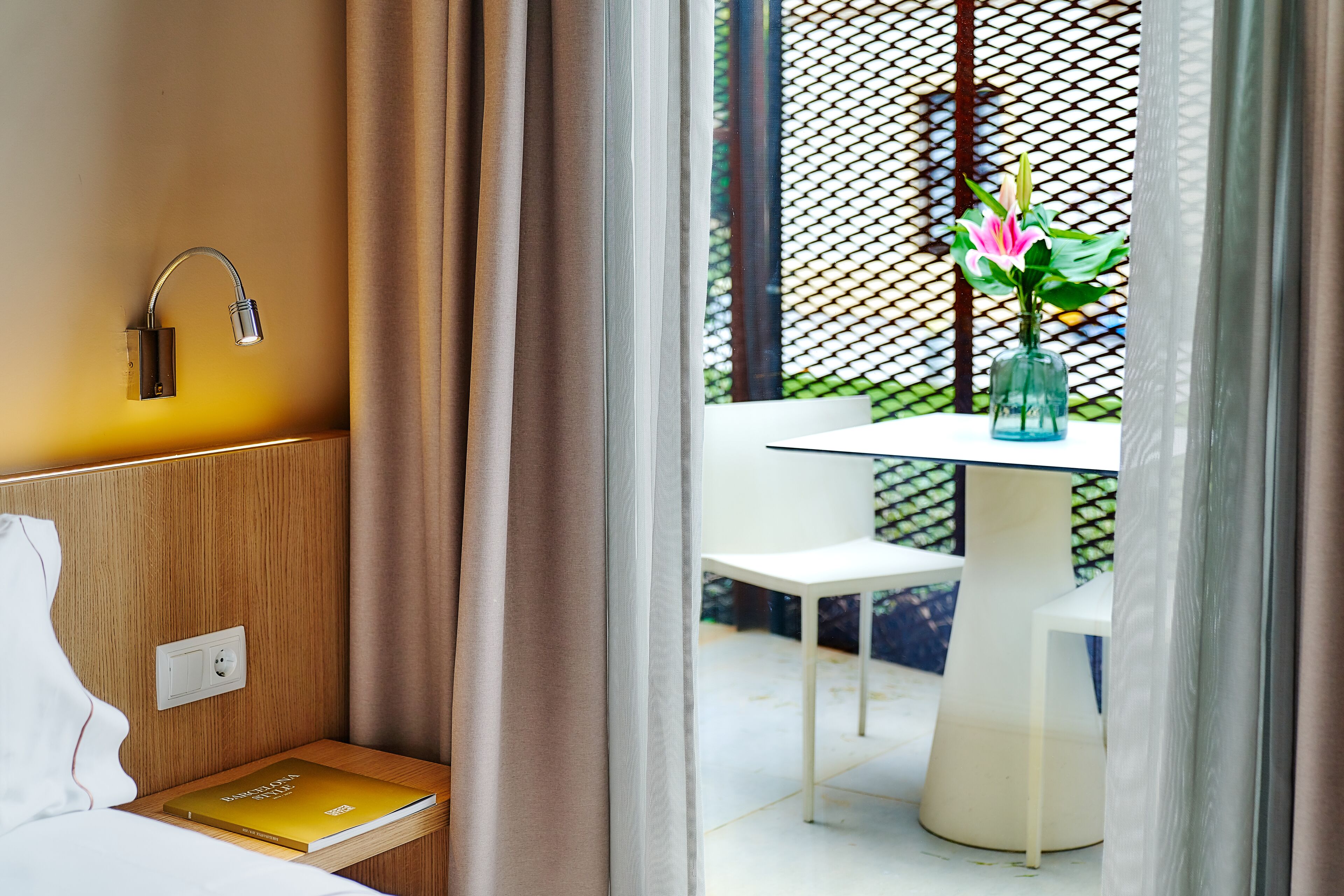 A hotel room with a bed, a bedside table with a book, a wall-mounted lamp, and a view of an outdoor seating area with a table and chairs.