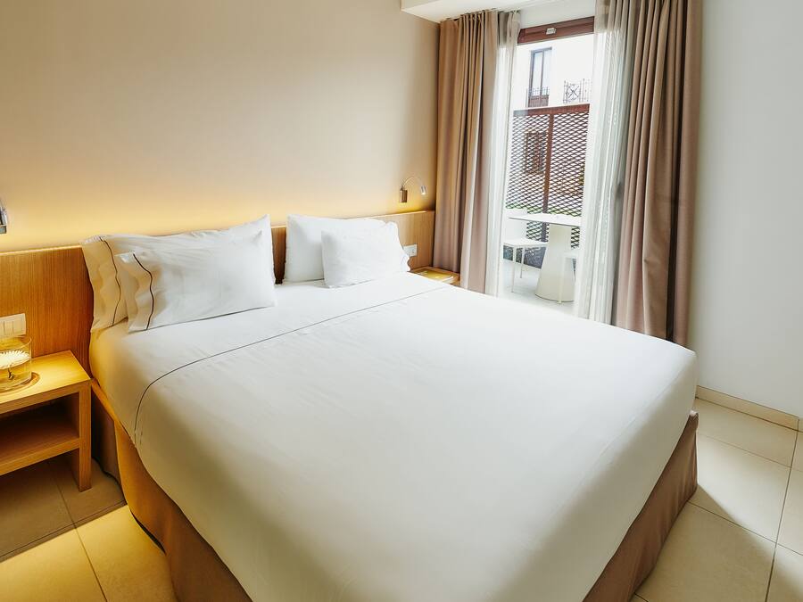 Una cama bien hecha con ropa blanca, una mesita de noche con lámpara y una ventana con cortinas.