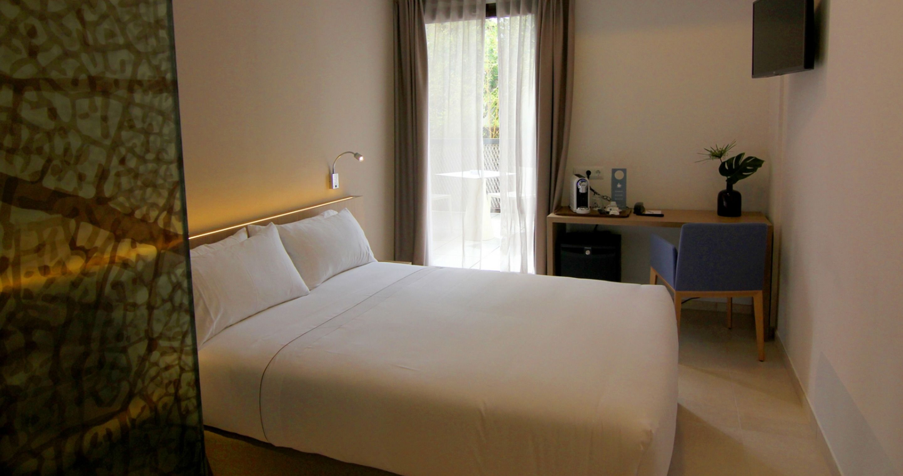 A hotel room with a large bed, a desk with a chair, a TV mounted on the wall, and a window with curtains.