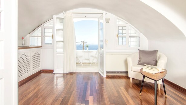 Amplia habitación con suelo de madera, techo abovedado blanco y vista al mar a través de un ventanal abierto.