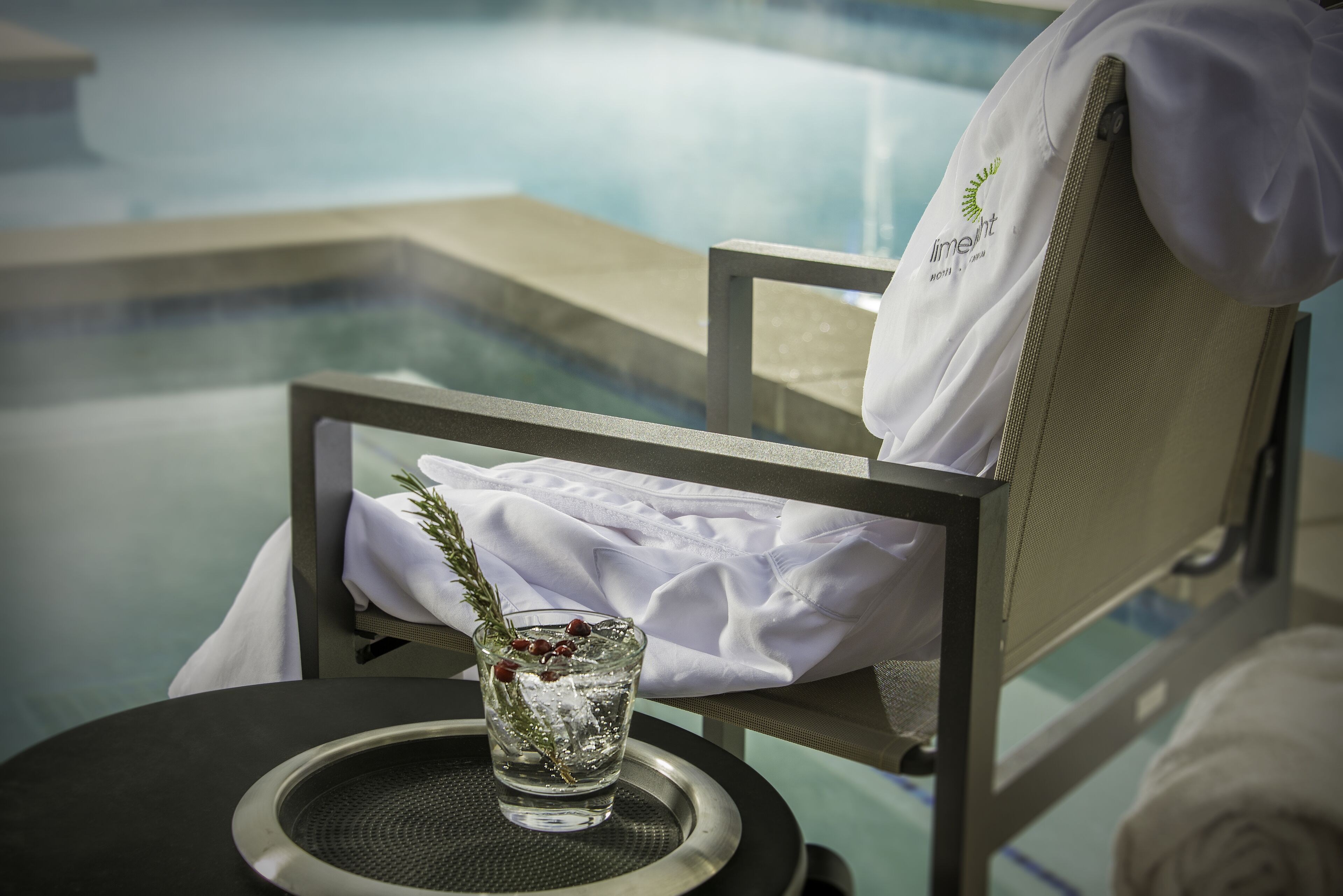 A chair by a pool with a glass of water and a sprig of rosemary.