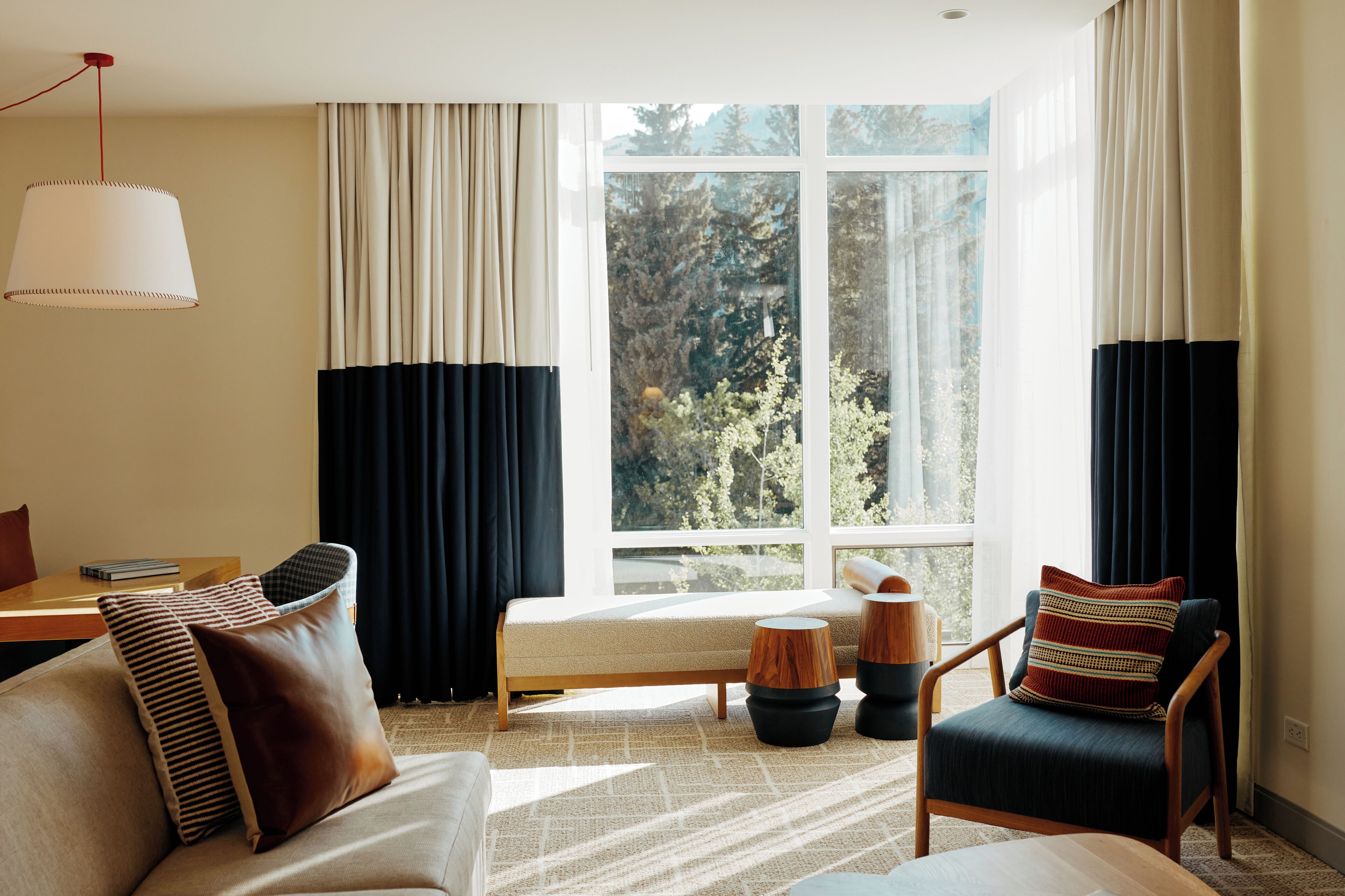 Una sala moderna con sofá, sillón y una mesita. Hay un ventanal con cortinas, y una lámpara cuelga del techo.