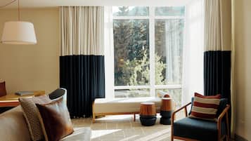 En moderne stue med sofa, lænestol og et lille bord. Der er et stort vindue med gardiner, og en lampe hænger fra loftet.