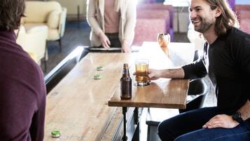 Una persona sentada en una mesa de madera con una botella y un vaso de cerveza, en un ambiente relajado con un sofá al fondo.