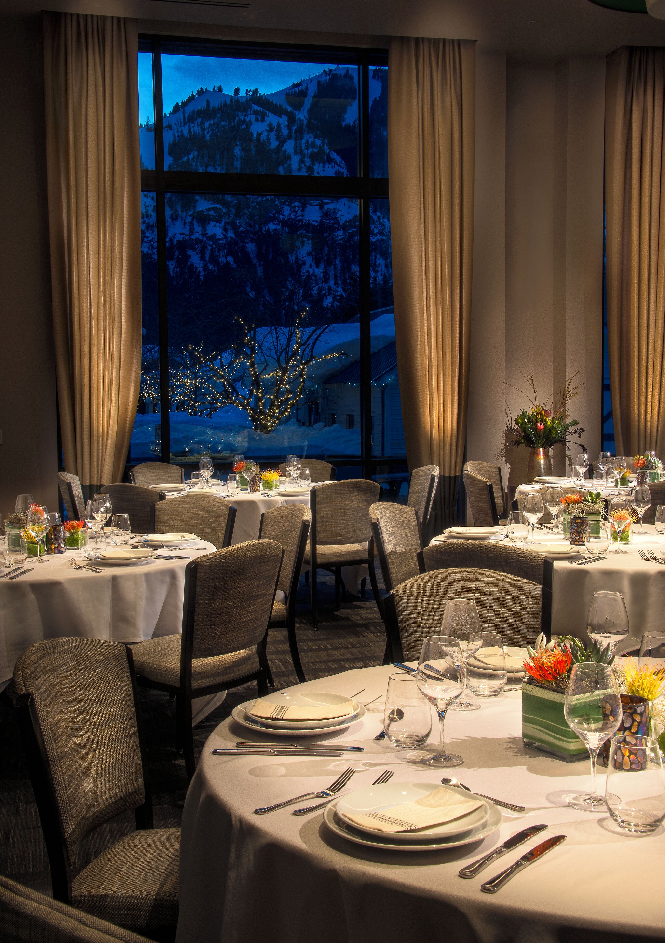 A restaurant with round tables set for dining, large windows with curtains, and a view of a snowy mountain.