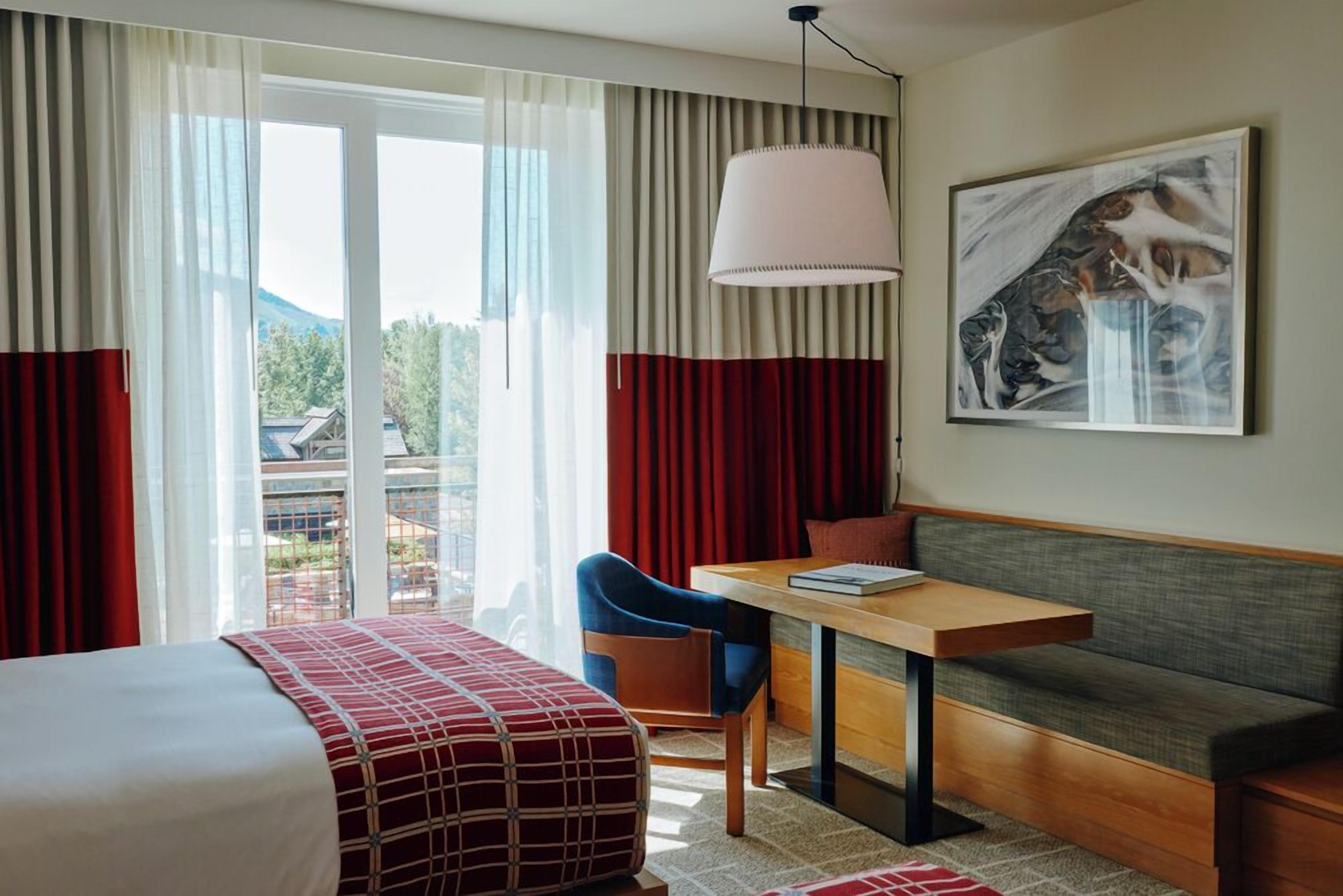 Habitación de hotel con cama, sofá, mesa y silla. Hay una ventana con cortinas, un cuadro en la pared y vistas al exterior.
