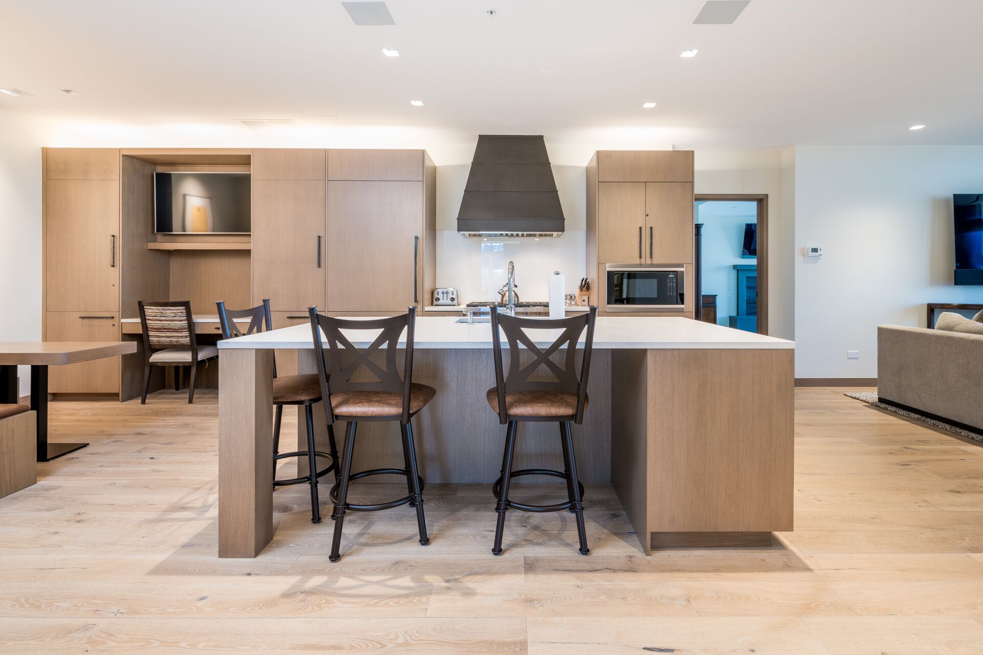 A modern kitchen with a central island, two bar stools, wooden cabinets, and a built-in microwave.