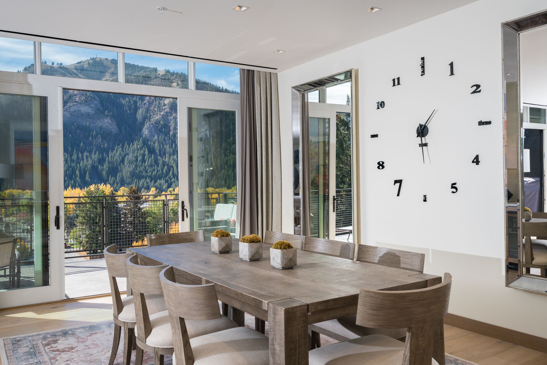 A dining area with a wooden table and chairs, large windows with mountain views, and a wall clock.
