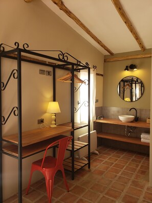 A compact room with a wooden desk, a red chair, a hanging clothes rack, a round mirror, and a bathroom with a sink and towel rack. - Casa de la Chola (Urubamba)