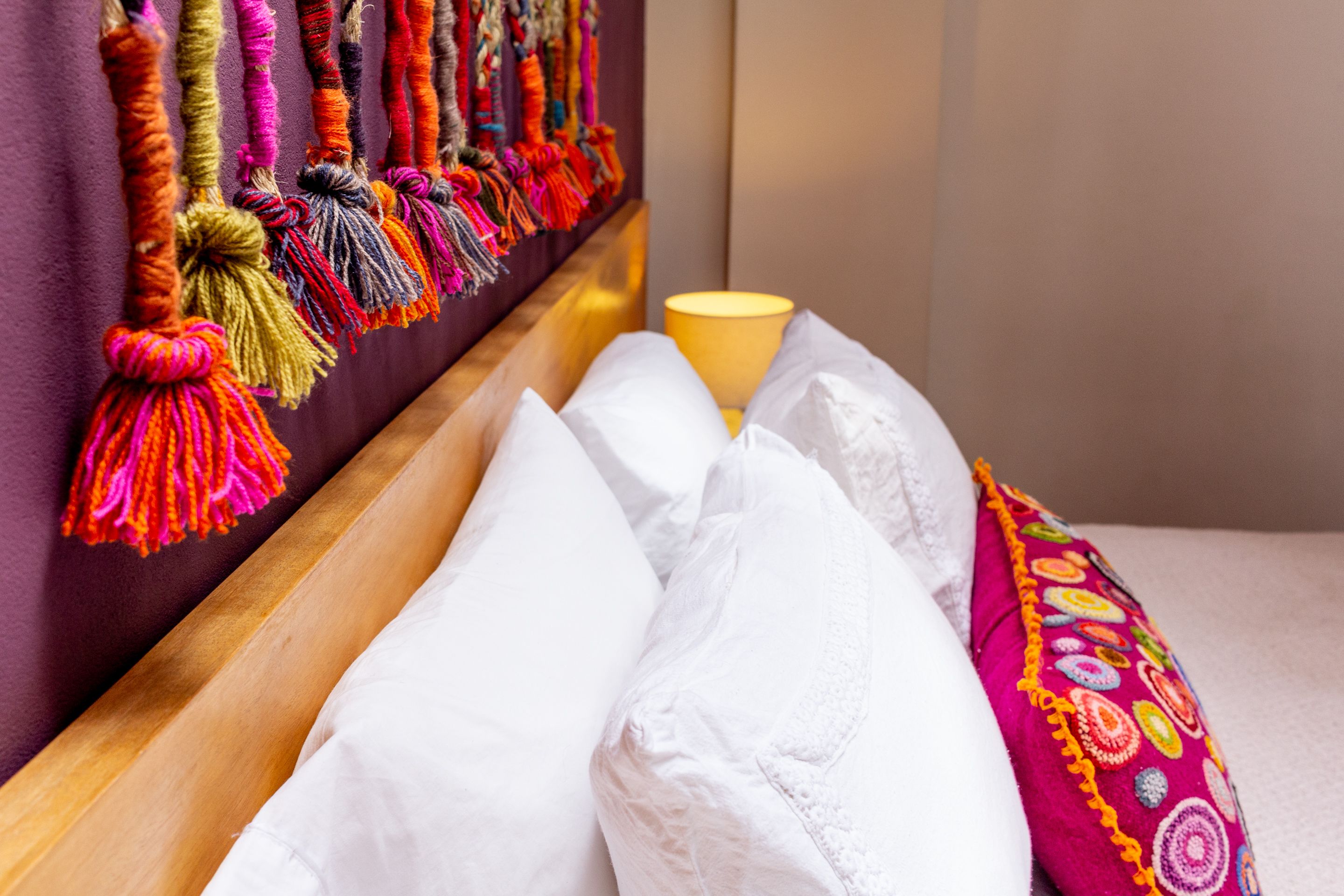 Una cama con almohadas blancas y una almohada bordada de colores, un cabecero de madera y una pared adornada con borlas colgantes.