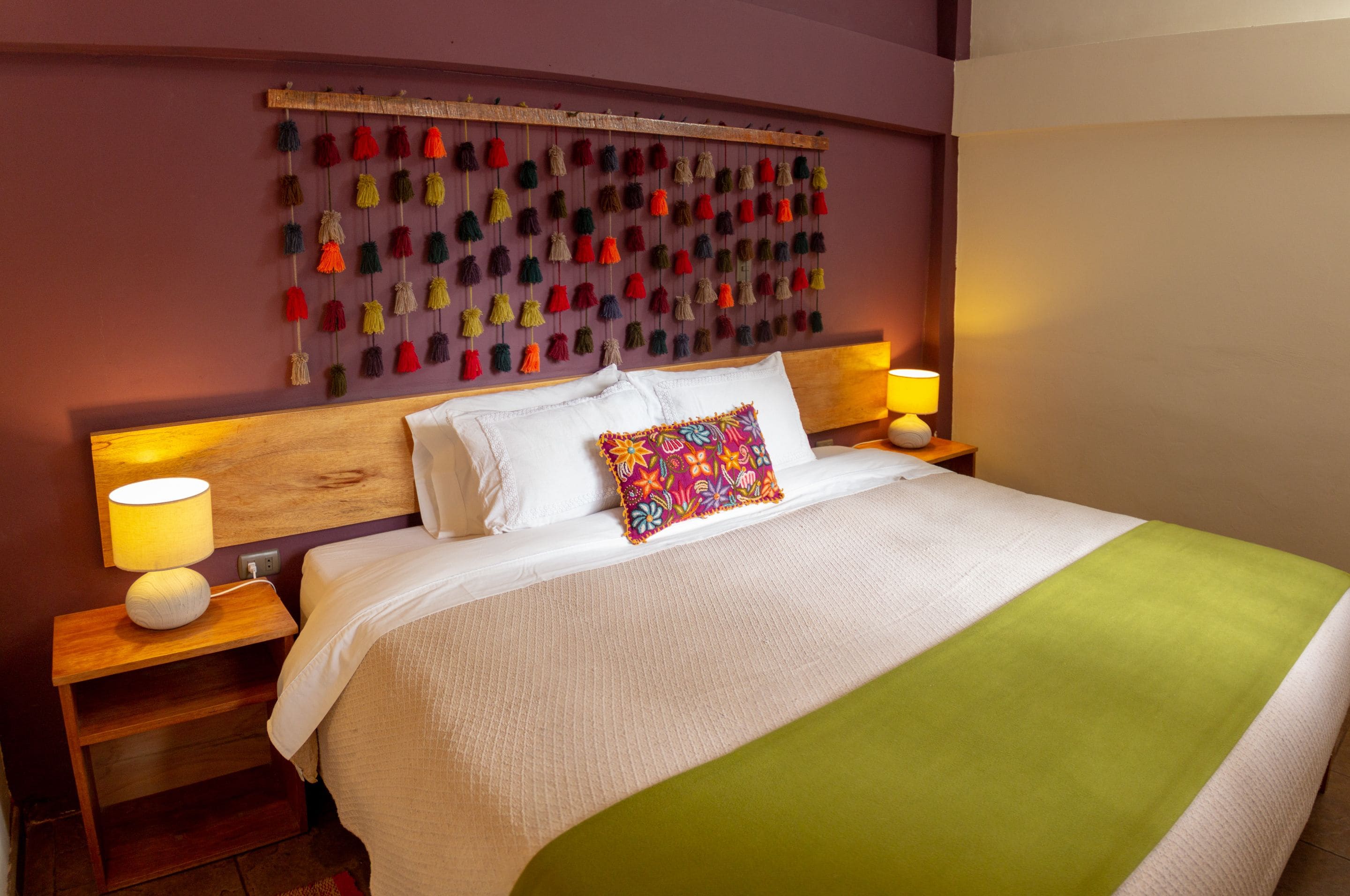 Una cama bien hecha con un cojín de colores, un cabecero con flecos colgantes y lámparas de noche sobre mesitas de madera.