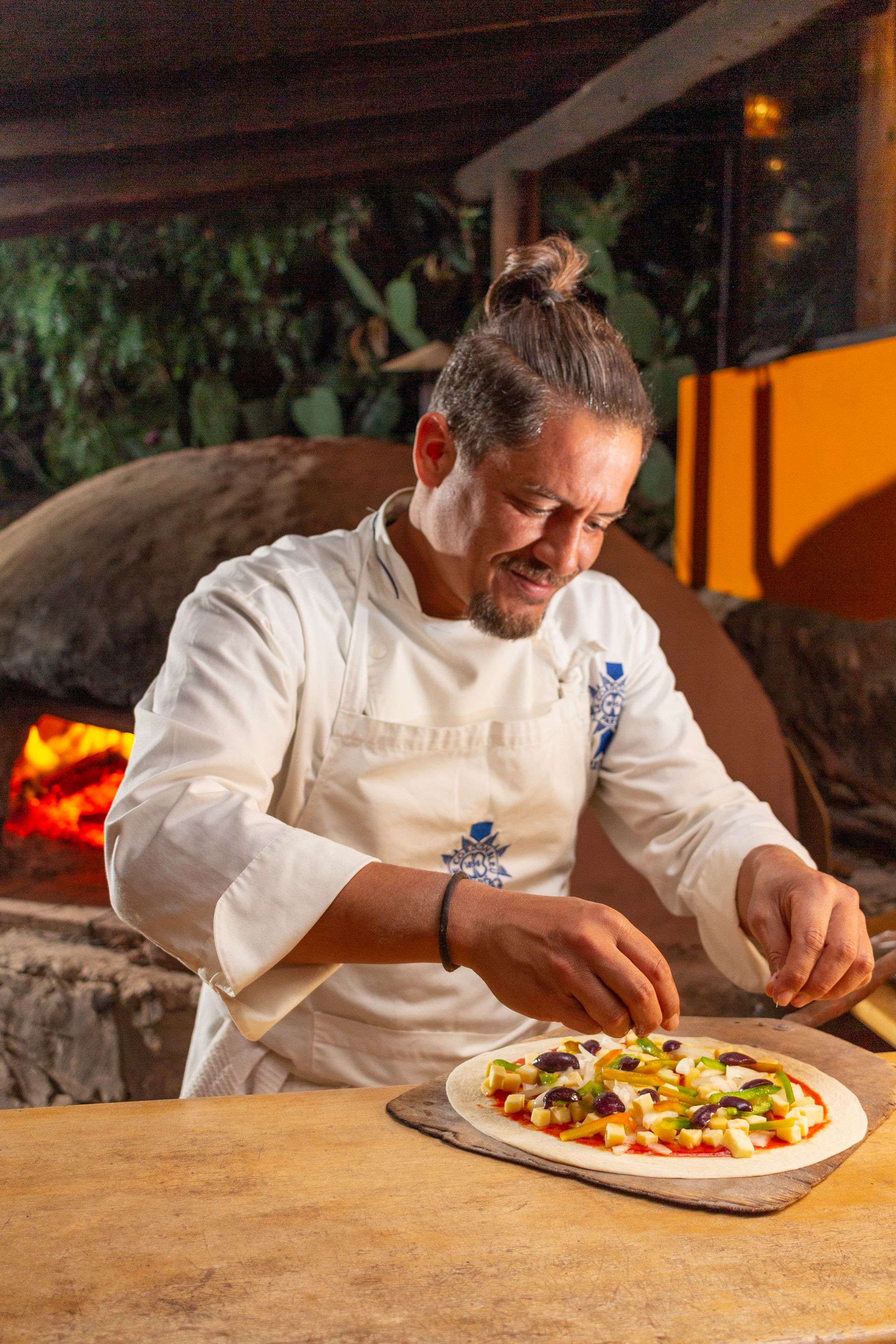 A chef preparing a pizza on a wooden board with a brick oven in the background.
