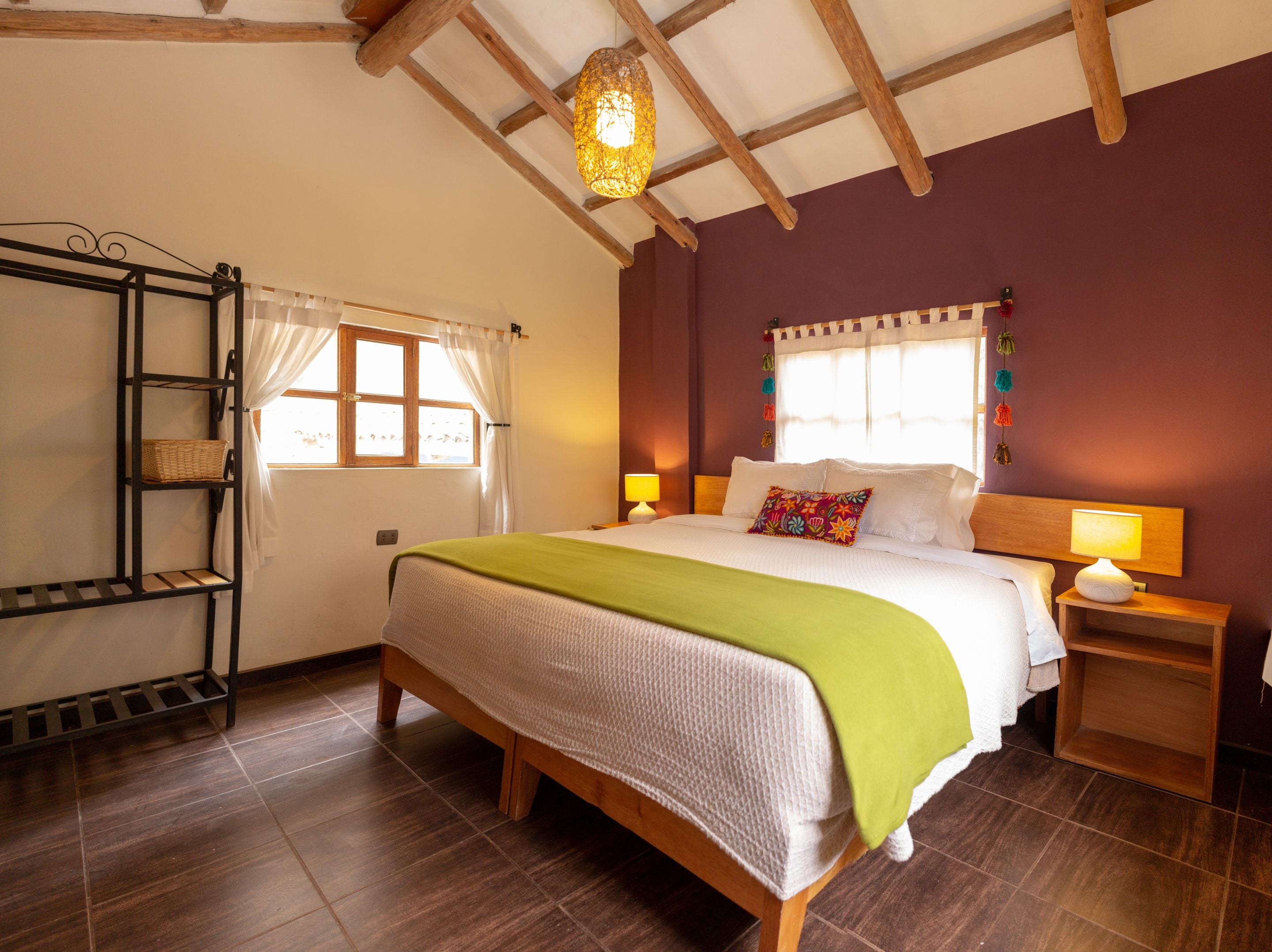 A bedroom with a wooden bed, a nightstand with a lamp, a window with curtains, and a shelf with a basket.