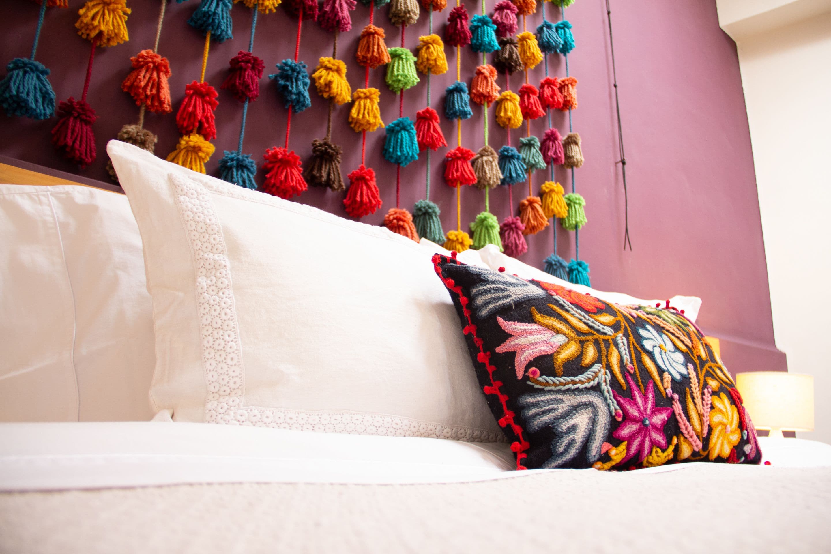 A bed with white linens and a colorful embroidered pillow against a purple wall with hanging tassels.